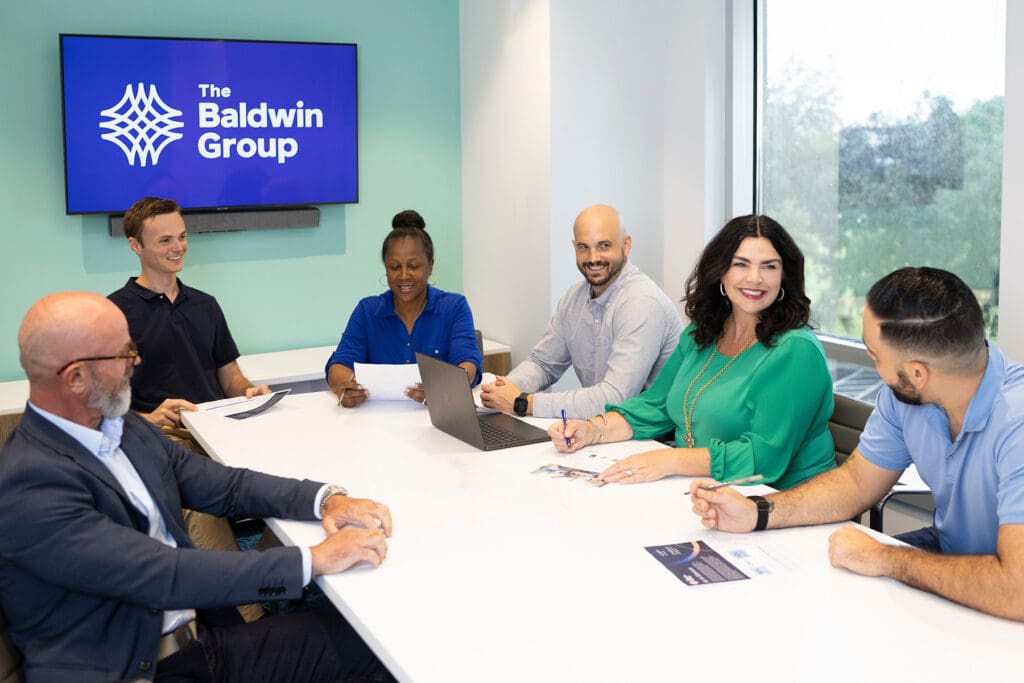 Colleagues meeting with founder partner in conference room - The Baldwin Group logo in background on TV