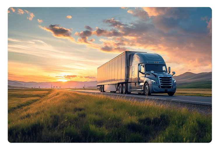 Low angle view of a gray semi truck with trailer driving on an asphalt road amidst green meadows. Summer trucking job during the sunset outdoors