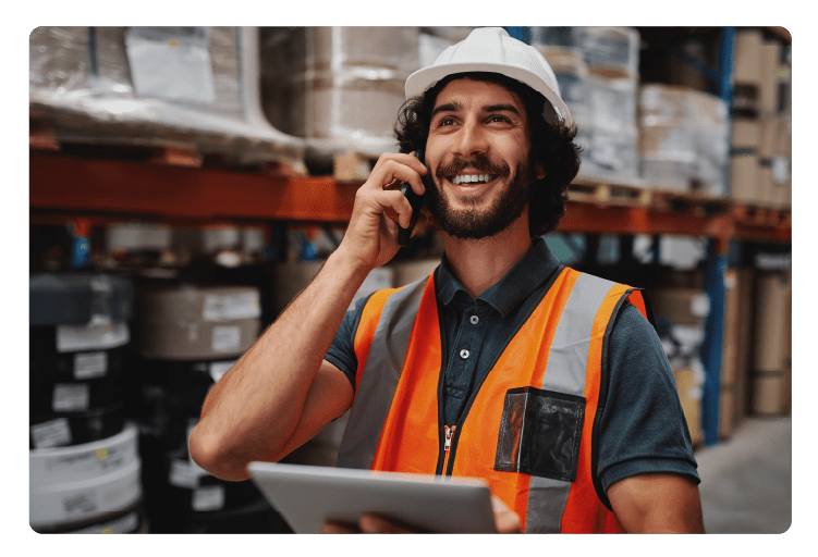 Warehouse worker using digital tablet while in conversation over mobile phone in warehouse wearing white helmet and safety vest