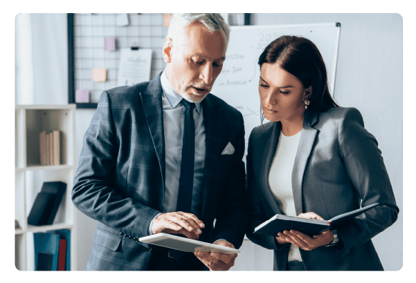 Mature investor using digital tablet near businesswoman with notebook in office