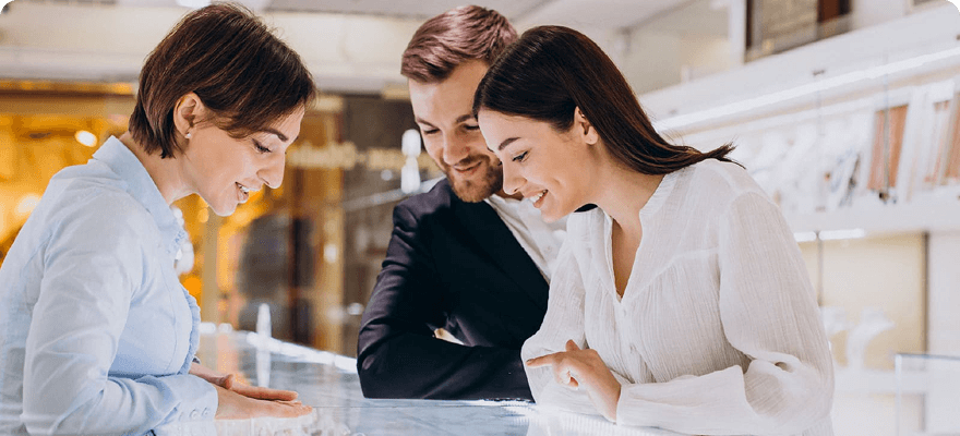 A man and a woman, both smiling, look at jewelry in a display case with the help of a store clerk. The clerk gestures toward the display