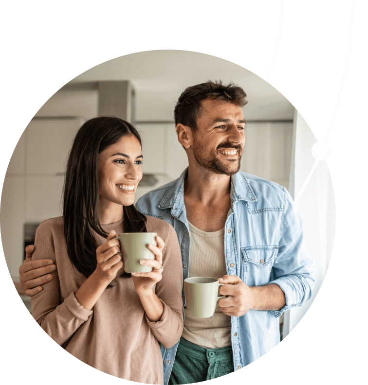 A smiling couple stands close together in a modern kitchen. The woman, on the left, holds a light-green mug with both hands, and the man, on the right, holds a similar mug and has his arm around the woman. The image is cropped into a circle.